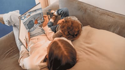 Betreuungsperson schaut gemeinsam mit einem Kind ein Bilderbuch auf dem Sofa an.