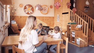 Pädagogische Fachkraft sitzt mit zwei Kindern an einem Tisch und nutzt ein Tablet zur Dokumentation im Betreuungsalltag.