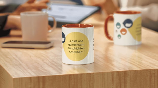 Kaffeetasse mit der Aufschrift „Lasst uns gemeinsam Geschichten schreiben“ steht auf einem Besprechungstisch, während sich Mitarbeitende im Hintergrund austauschen.