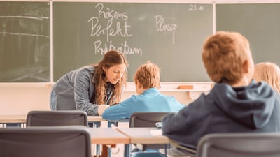 Lehrkraft unterstützt ein Grundschulkind beim Lernen im Klassenzimmer, während weitere Kinder an ihren Tischen sitzen.