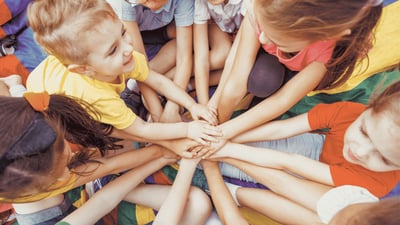 Mehrere Kinder sitzen im Kreis und legen ihre Hände übereinander als Zeichen für Gemeinschaft und Zusammenhalt.