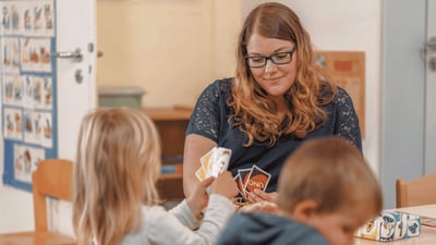 Pädagogische Fachkraft spielt mit zwei Kindern an einem Tisch und begleitet sie beim gemeinsamen Kartenspiel.