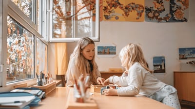 Pädagogische Fachkraft unterstützt ein Kind beim gemeinsamen Spielen an einem Tisch in einer Betreuungseinrichtung.