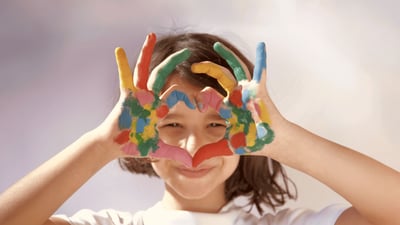 Lachendes Kind hält seine bunt bemalten Hände vor das Gesicht und formt mit den Fingern eine Brille.